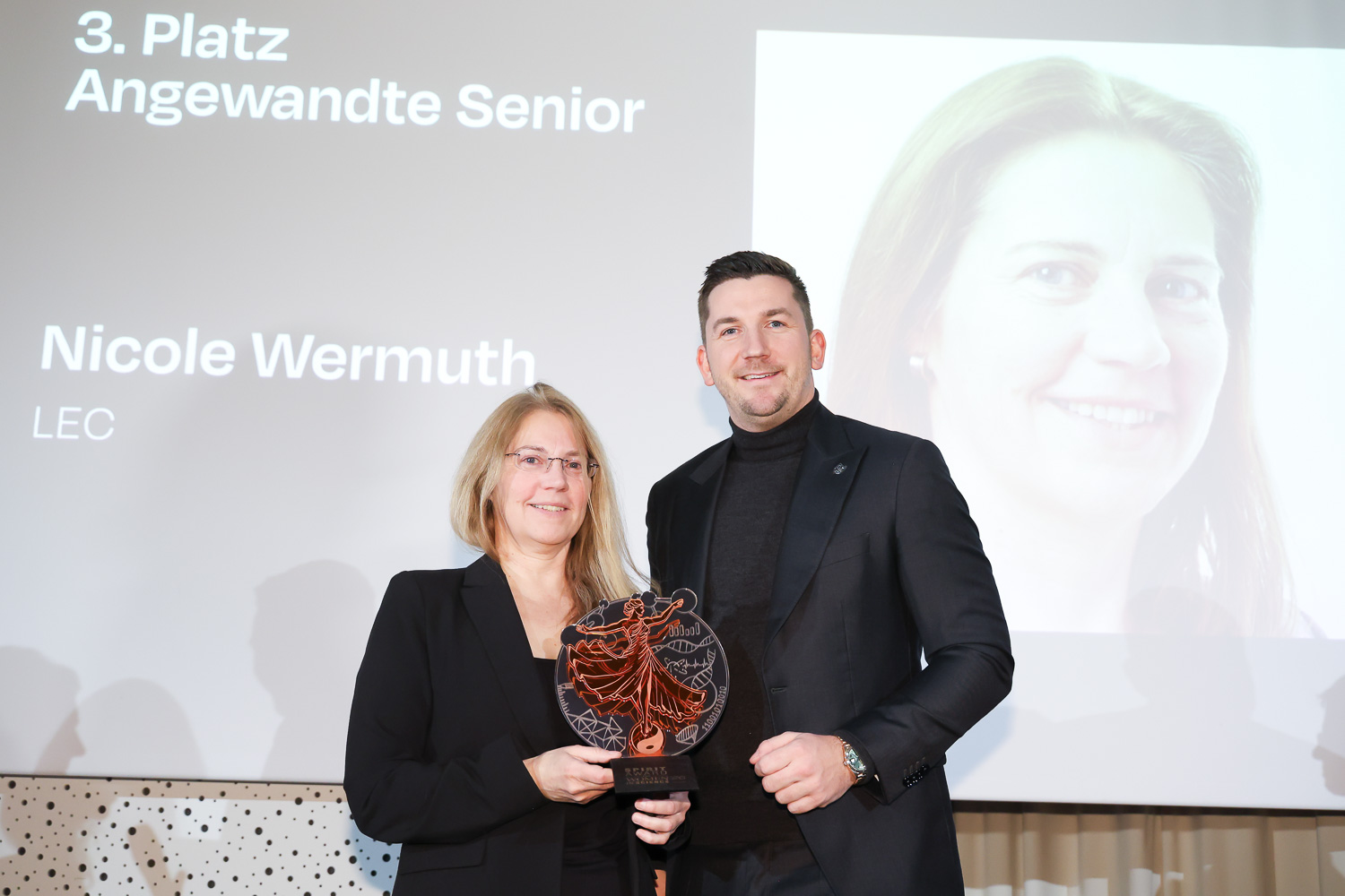 Zwei formell gekleidete Personen halten auf der Bühne einen Preis, hinter ihnen wird auf einer Leinwand "Nicole Wermuth" und "3. Platz Angewandte Senior" angezeigt.