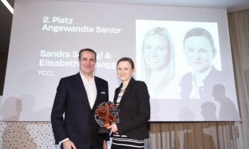 Ein Mann und eine Frau halten lächelnd eine Trophäe auf der Bühne, während ihre Namen und Porträts auf einem Bildschirm hinter ihnen bei einer Preisverleihung angezeigt werden.