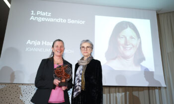 Zwei Frauen stehen lächelnd bei einer Preisverleihung; eine hält eine Trophäe. Auf einem Bildschirm hinter ihnen ist "1. Platz Angewandte Senior" und ein Porträtfoto zu sehen.