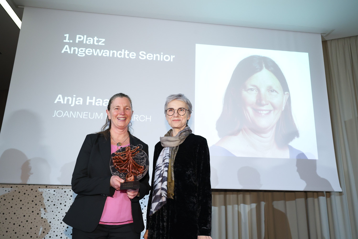 Zwei Frauen stehen lächelnd bei einer Preisverleihung; eine hält eine Trophäe. Auf einem Bildschirm hinter ihnen ist "1. Platz Angewandte Senior" und ein Porträtfoto zu sehen.