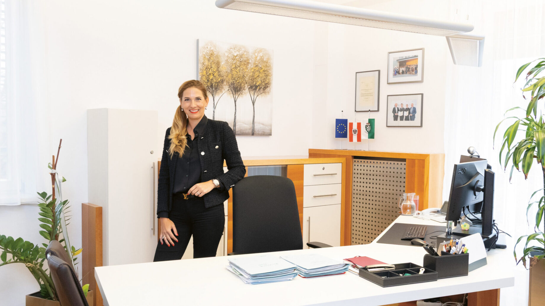 Eine Frau steht lächelnd in einem modernen, hellen Büro mit einem Schreibtisch, einem Computer, Pflanzen und gerahmten Kunstwerken an den Wänden.