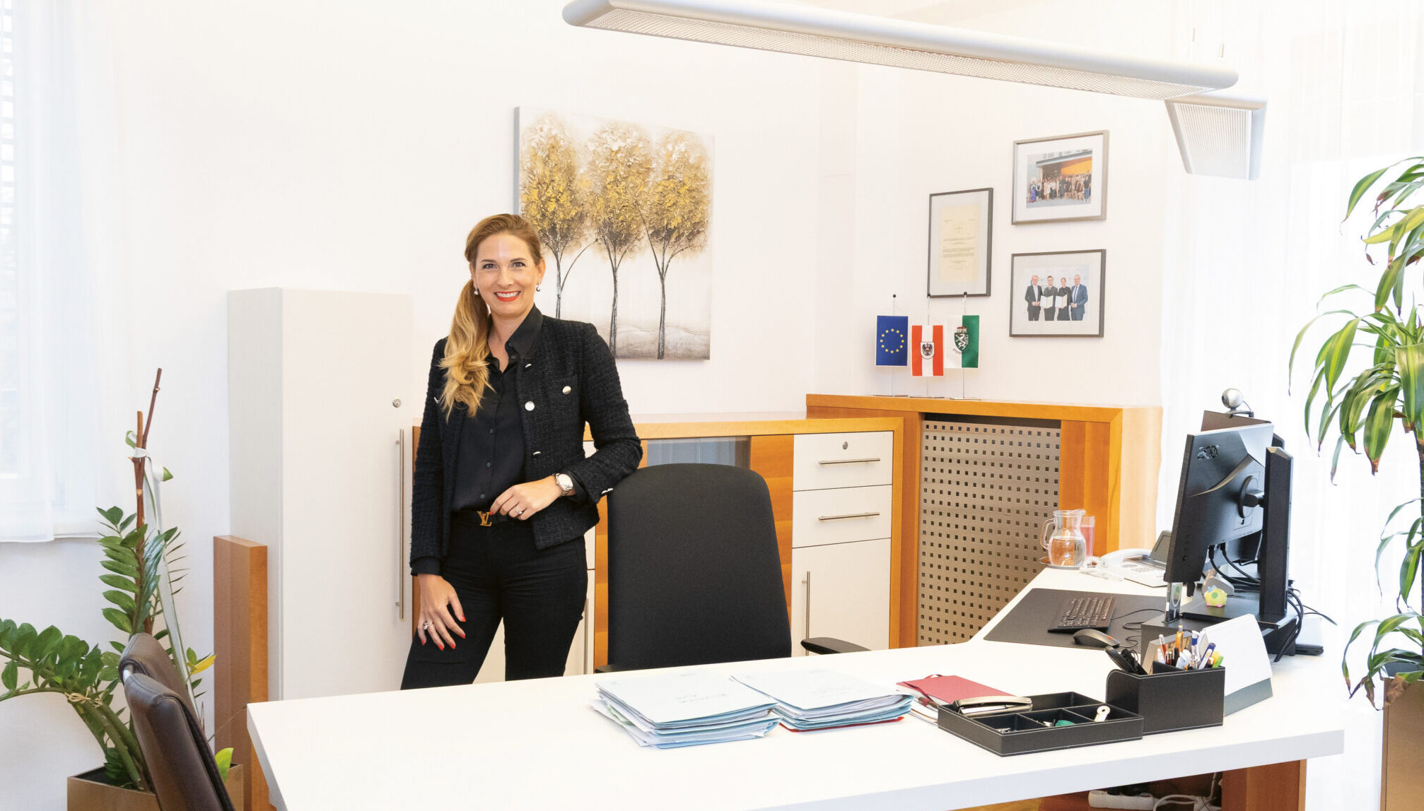 Eine Frau steht lächelnd in einem modernen, hellen Büro mit einem Schreibtisch, einem Computer, Pflanzen und gerahmten Kunstwerken an den Wänden.