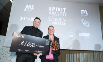Zwei lächelnde Personen, die einen großen Scheck über 1 000 Euro und eine Trophäe bei der Verleihung des Spirit Award for Women in Science in der Hand halten, dahinter ein Bildschirm.