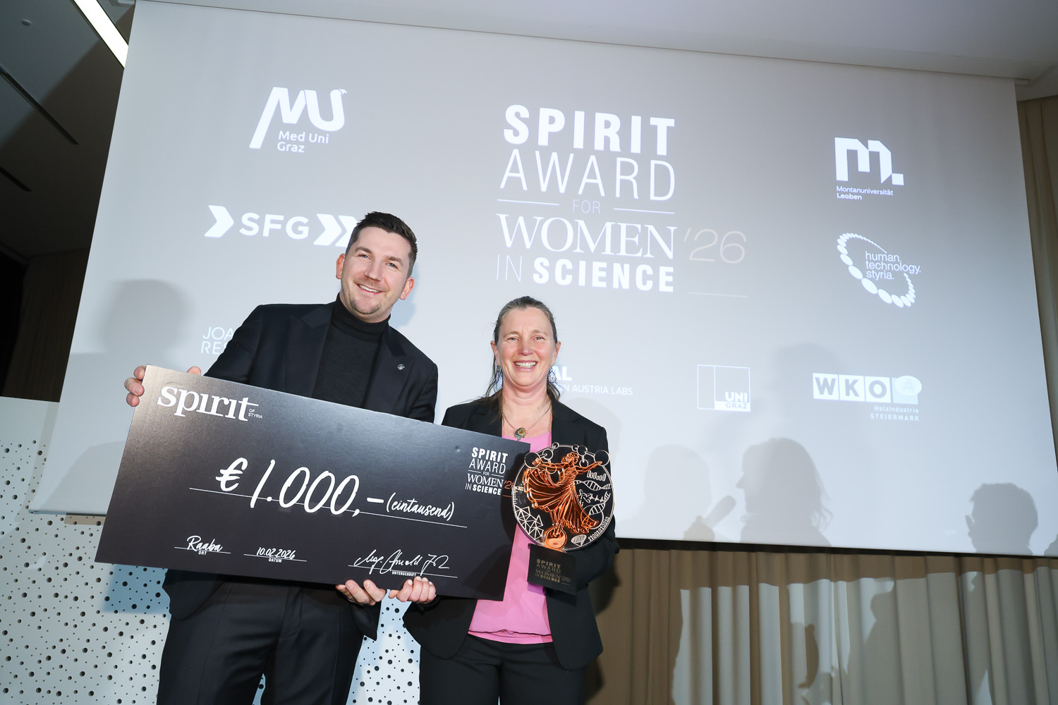 Zwei lächelnde Personen, die einen großen Scheck über 1 000 Euro und eine Trophäe bei der Verleihung des Spirit Award for Women in Science in der Hand halten, dahinter ein Bildschirm.