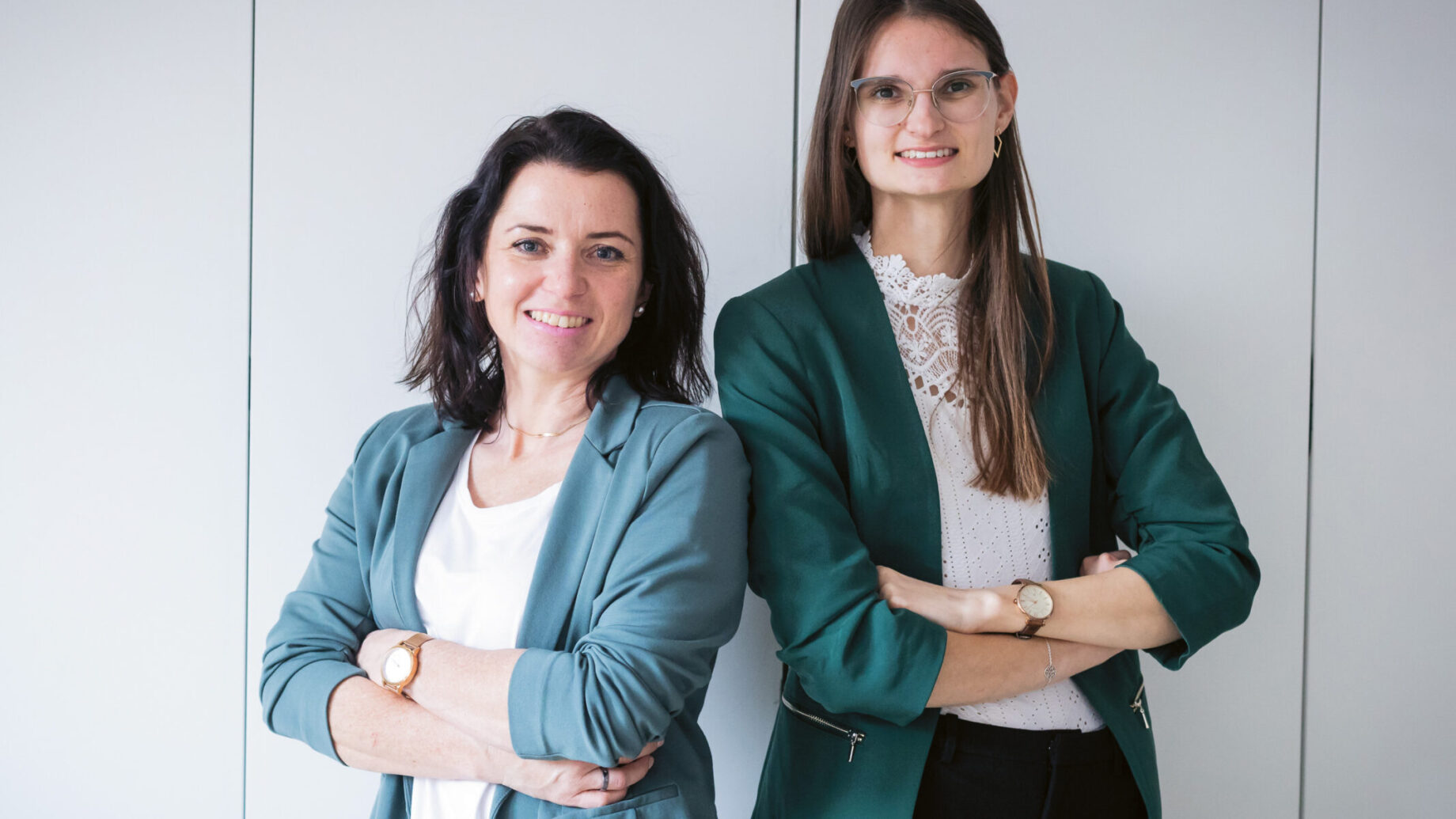 Zwei Frauen in Geschäftskleidung stehen mit verschränkten Armen nebeneinander und lächeln vor einem schlichten hellen Hintergrund.