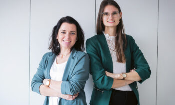 Zwei Frauen in Geschäftskleidung stehen mit verschränkten Armen nebeneinander und lächeln vor einem schlichten hellen Hintergrund.