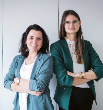 Zwei Frauen in Geschäftskleidung stehen mit verschränkten Armen nebeneinander und lächeln vor einem schlichten hellen Hintergrund.