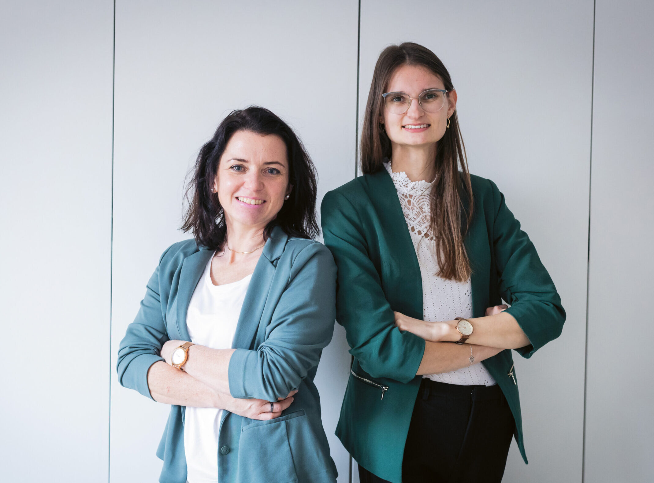 Zwei Frauen in Geschäftskleidung stehen mit verschränkten Armen nebeneinander und lächeln vor einem schlichten hellen Hintergrund.