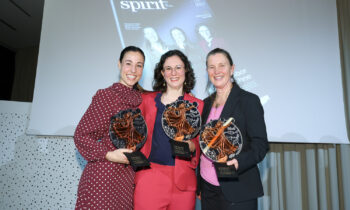 Drei lächelnde Frauen stehen zusammen, halten Trophäen in der Hand und posieren vor einem projizierten Bild eines Magazintitels mit der Aufschrift "spirit".