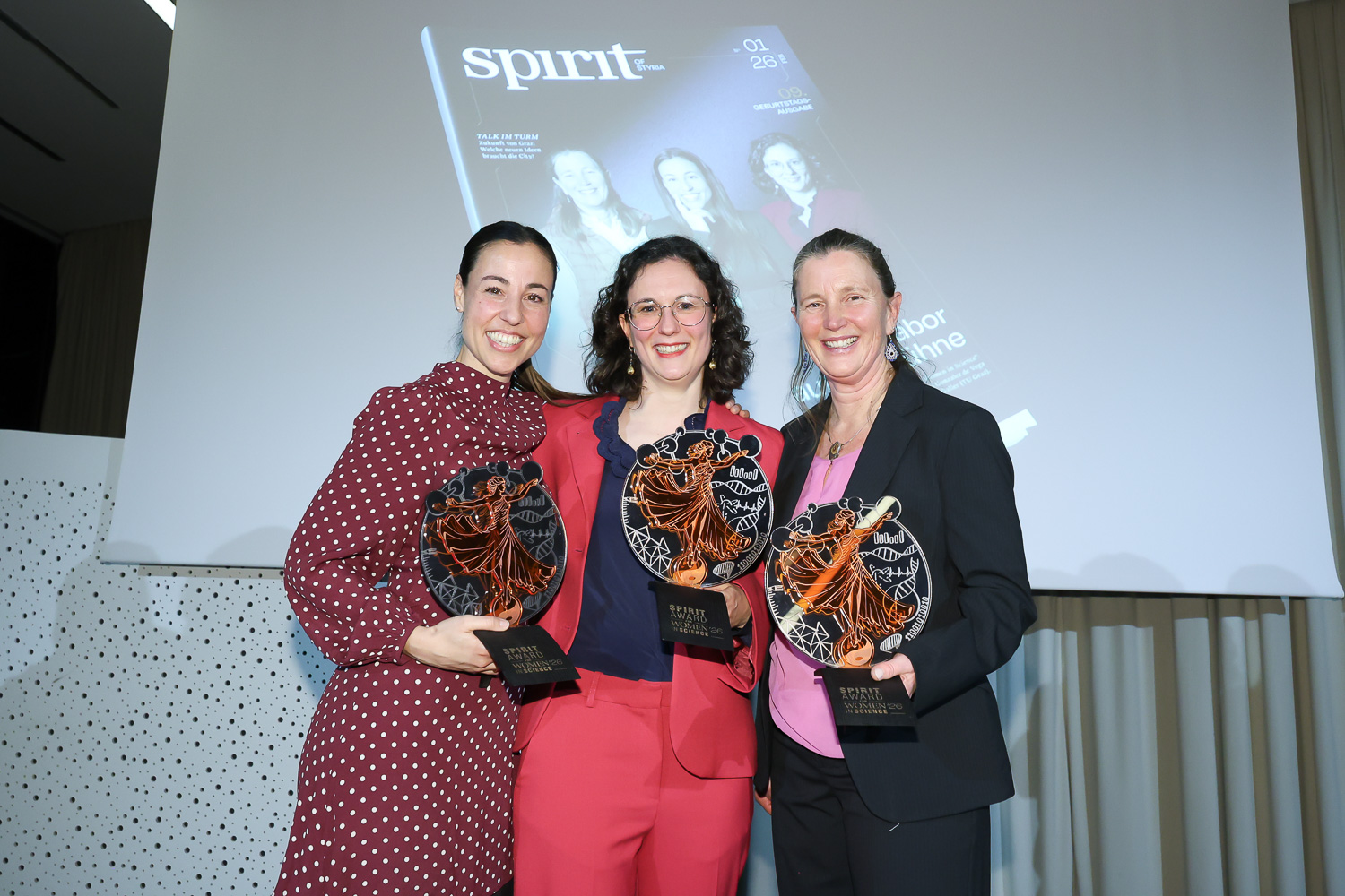 Drei lächelnde Frauen stehen zusammen, halten Trophäen in der Hand und posieren vor einem projizierten Bild eines Magazintitels mit der Aufschrift "spirit".