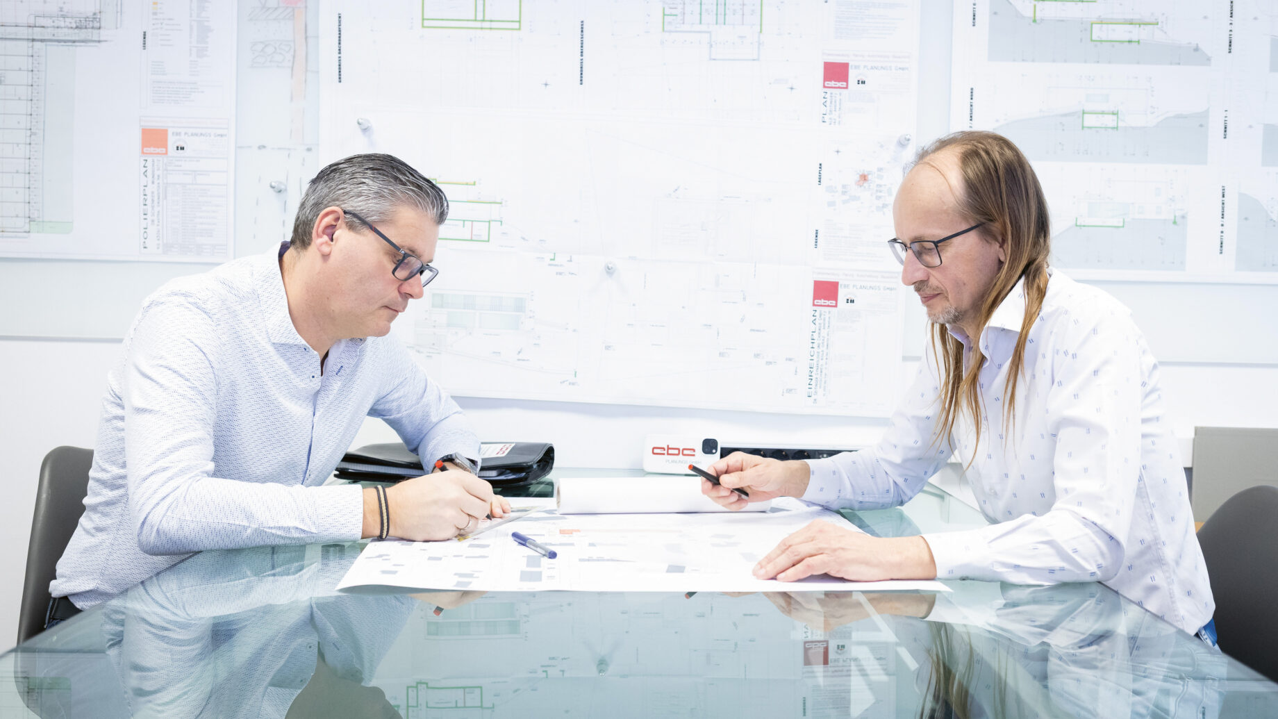 Zwei Männer sitzen in einem hellen Büro an einem Glastisch und besprechen Architekturpläne mit Blaupausen und Diagrammen an der Wand hinter ihnen.