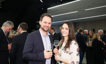 Ein Mann und eine Frau lächeln und halten einen Drink bei einer formellen Veranstaltung, während sich im Hintergrund andere Leute treffen.