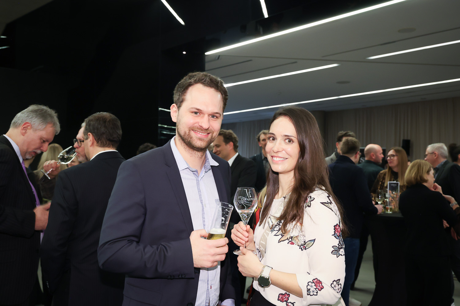 Ein Mann und eine Frau lächeln und halten einen Drink bei einer formellen Veranstaltung, während sich im Hintergrund andere Leute treffen.