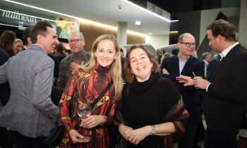 Zwei Frauen lächeln bei einem gesellschaftlichen Ereignis, sie halten Getränke in der Hand, im Hintergrund unterhalten sich mehrere Personen in einer modernen Innenumgebung.