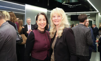 Zwei Frauen lächeln und posieren zusammen bei einer formellen Veranstaltung in einem Gebäude, während sich im Hintergrund andere Menschen treffen.