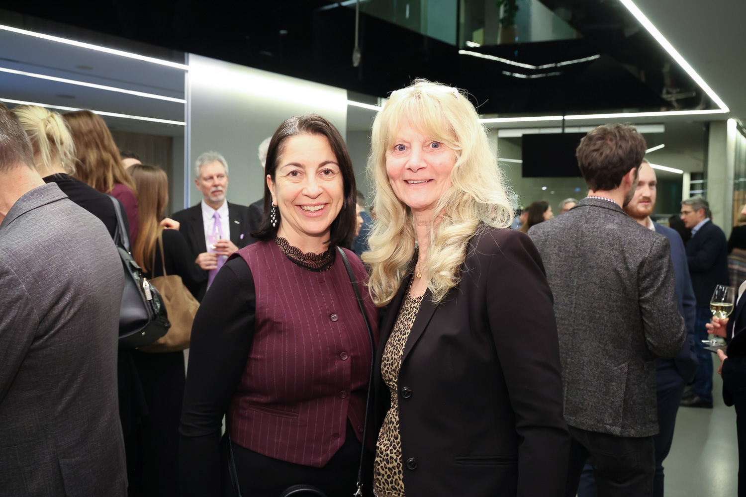 Zwei Frauen lächeln und posieren zusammen bei einer formellen Veranstaltung in einem Gebäude, während sich im Hintergrund andere Menschen treffen.