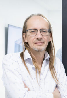 Zwei Männer in hellen Hemden stehen in einem hellen, modernen Büro, einer mit Brille und kurzen Haaren, der andere mit langen Haaren und verschränkten Armen.