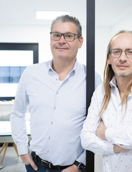 Zwei Männer in hellen Hemden stehen nebeneinander in einem hellen, modernen Büro, einer mit Brille, der andere mit langen Haaren und verschränkten Armen.