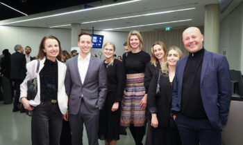 Sieben Personen in Geschäftskleidung posieren und lächeln gemeinsam bei einer Veranstaltung oder Konferenz in einem Gebäude, während sich andere im Hintergrund unter sie mischen.