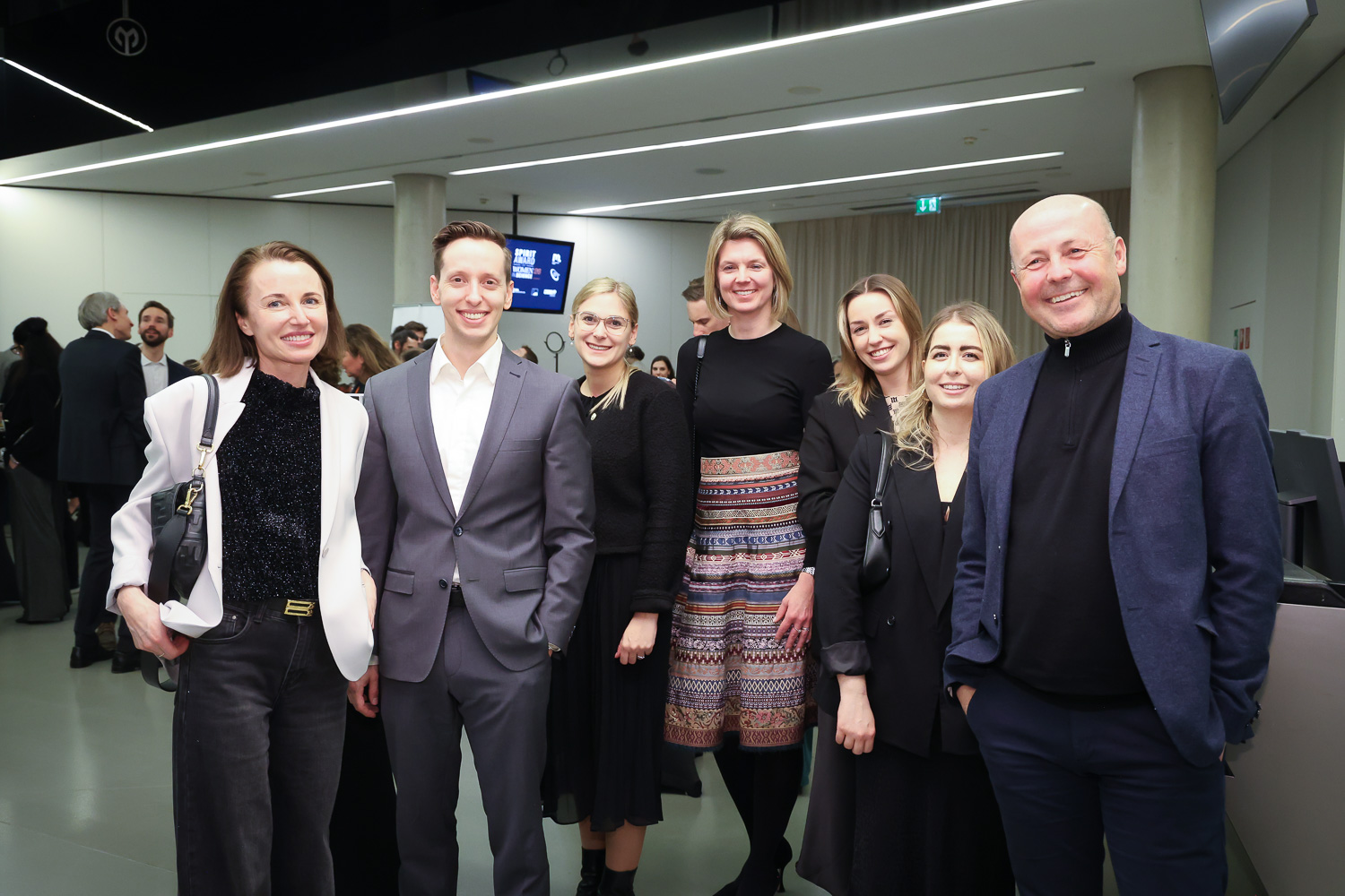 Sieben Personen in Geschäftskleidung posieren und lächeln gemeinsam bei einer Veranstaltung oder Konferenz in einem Gebäude, während sich andere im Hintergrund unter sie mischen.