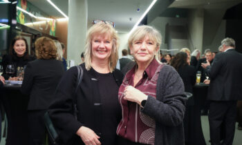 Zwei Frauen lächeln und posieren zusammen bei einer formellen Veranstaltung in einem Gebäude mit Menschen, die sich im Hintergrund treffen.