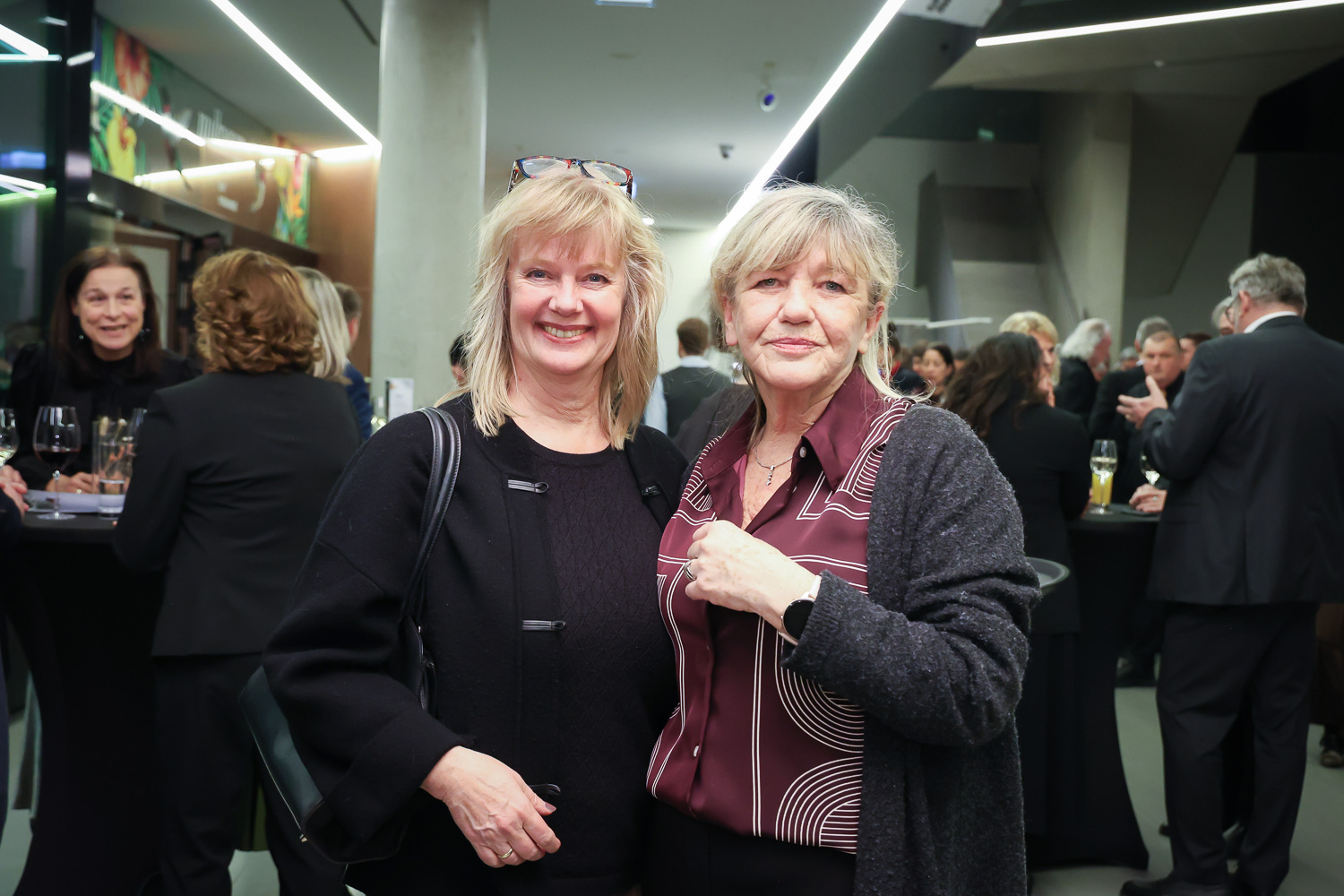 Zwei Frauen lächeln und posieren zusammen bei einer formellen Veranstaltung in einem Gebäude mit Menschen, die sich im Hintergrund treffen.