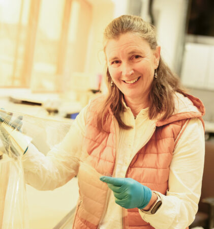 Eine lächelnde Frau in einem Laborkittel und mit Handschuhen hält eine Klarsichtfolie in einer Laborumgebung und steht neben großen Filmrollen auf einem Tisch.