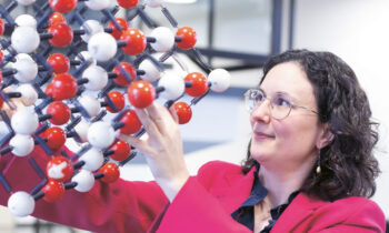 Eine Frau in einem leuchtend rosafarbenen Blazer untersucht ein großes Molekularmodell aus roten, weißen und schwarzen Kugeln in einem Raum.