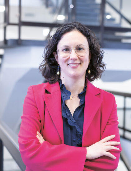 Eine Frau mit lockigem Haar und Brille, die einen rosa Blazer trägt, steht lächelnd mit verschränkten Armen vor einer modernen Treppe.
