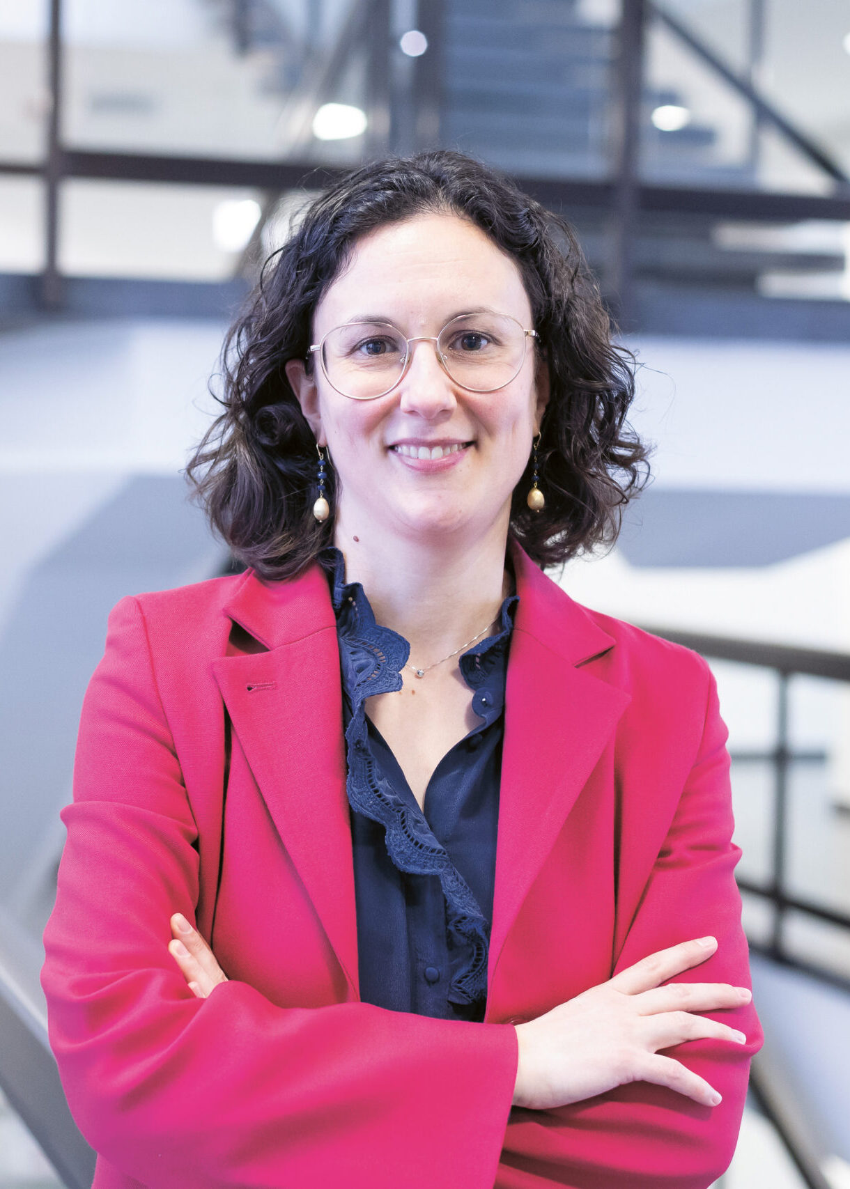 Frau mit lockigem Haar, Brille und rosa Blazer steht lächelnd mit verschränkten Armen in einem modernen Gebäude mit Treppen im Hintergrund.