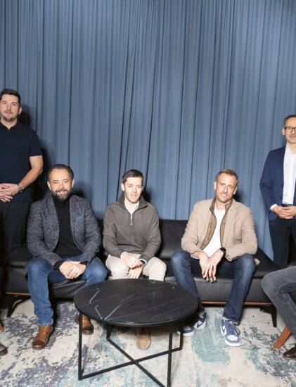 Sieben Personen posieren gemeinsam in einem Innenraum, sitzend und stehend vor blauen Vorhängen, mit einem "Geist"-Schild im Hintergrund.