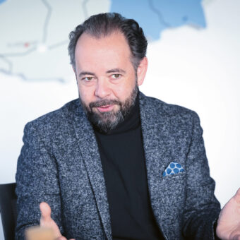Ein Mann mit Bart und Schnurrbart gestikuliert, während er spricht. Er trägt einen grauen Blazer und einen schwarzen Rollkragenpullover und sitzt vor einer Landkarte an der Wand.