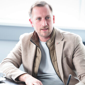 Ein Mann in einer beigen Jacke sitzt an einem Schreibtisch mit einem Tablet und einem Stift, einer Kaffeetasse und Papieren vor sich und scheint ein Gespräch zu führen.
