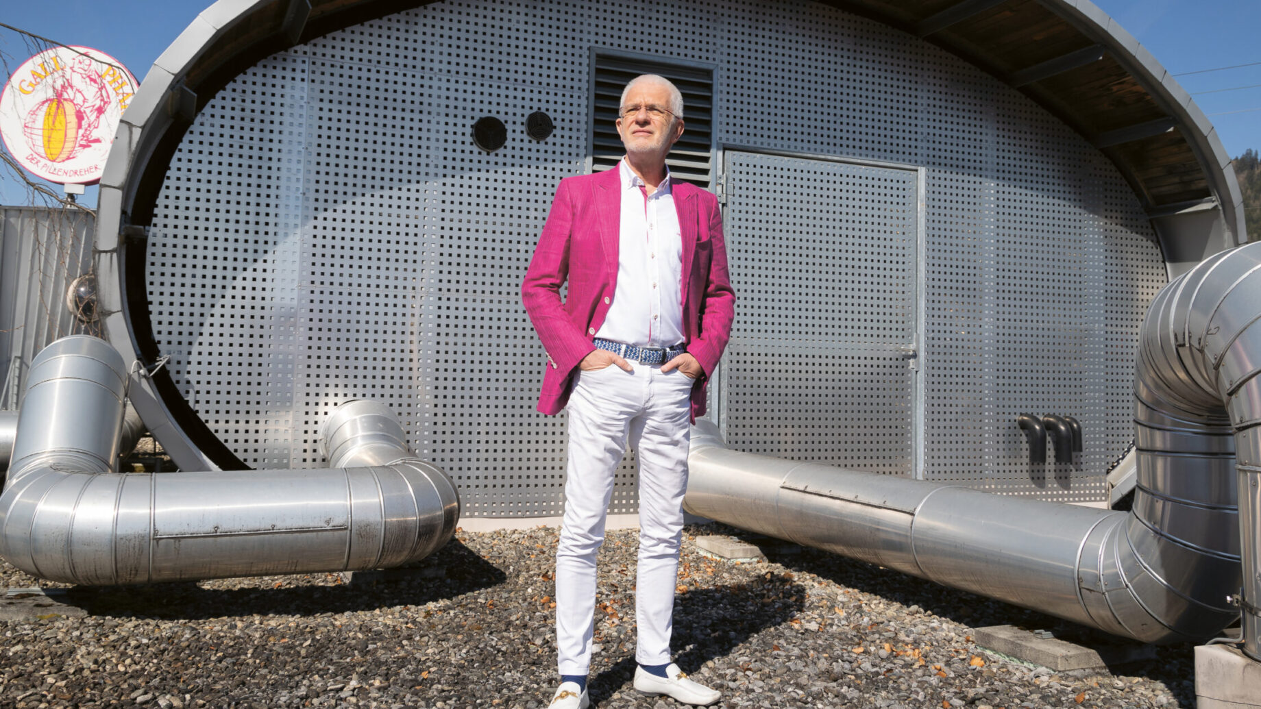 Ein Mann in einem leuchtend rosa Blazer steht vor einem modernen, metallischen Gebäude mit Rohren und einem sonnigen Himmel über ihm.