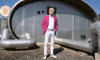 Ein Mann in einem leuchtend rosa Blazer steht vor einem modernen, metallischen Gebäude mit Rohren und einem sonnigen Himmel über ihm.
