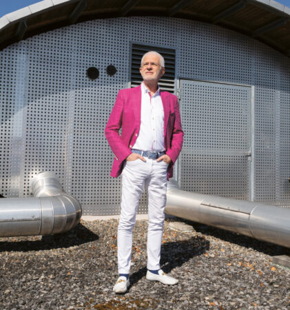 Ein Mann in einem leuchtend rosa Blazer steht vor einem modernen, metallischen Gebäude mit Rohren und einem sonnigen Himmel über ihm.