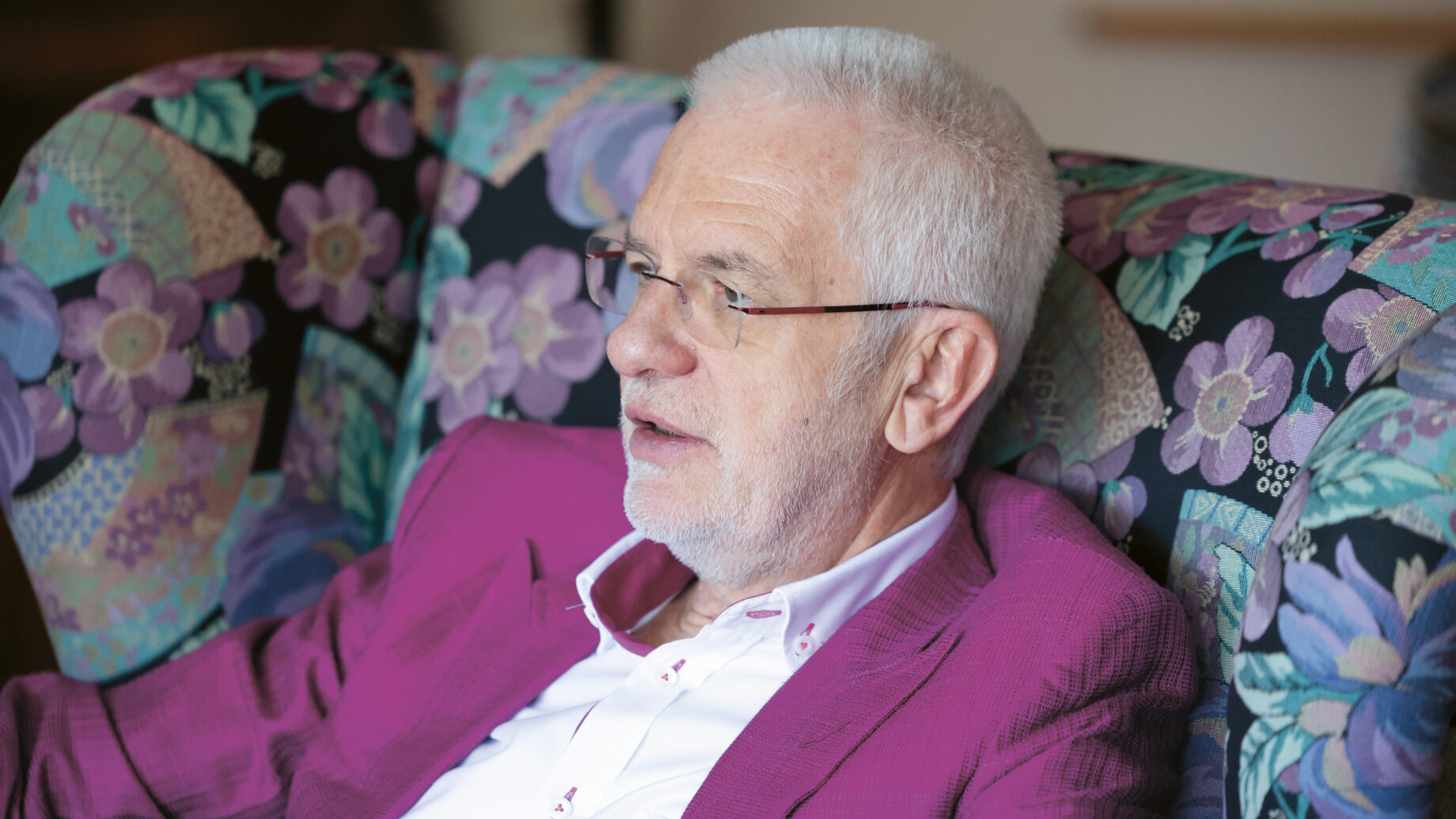 Ein älterer Mann mit weißem Haar und Brille, der eine magentafarbene Jacke trägt, sitzt auf einem geblümten Stuhl und schaut zur Seite.