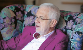 Ein älterer Mann mit weißem Haar und Brille, der eine magentafarbene Jacke trägt, sitzt auf einem geblümten Stuhl und schaut zur Seite.