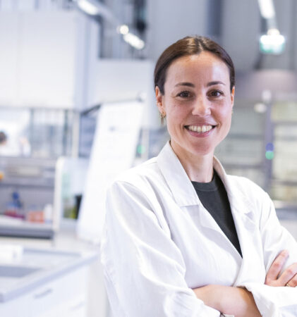 Ein lächelnder Wissenschaftler in einem weißen Laborkittel steht mit verschränkten Armen in einem hellen, modernen Labor.