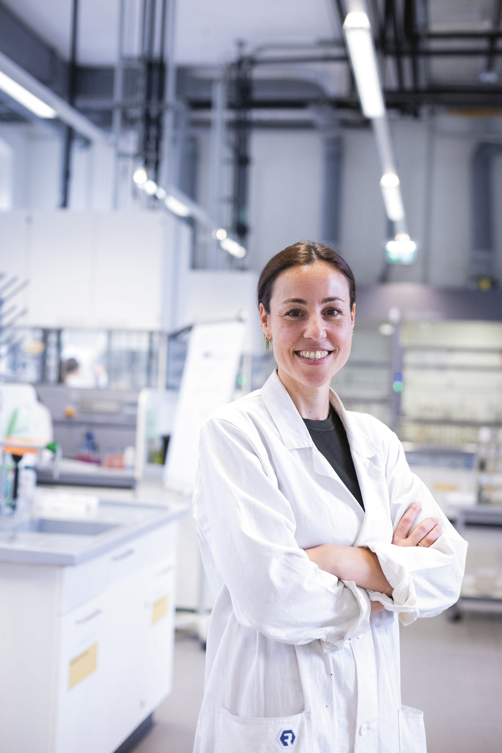 Eine Frau in einem weißen Laborkittel steht lächelnd mit verschränkten Armen in einer hellen, modernen Laborumgebung.