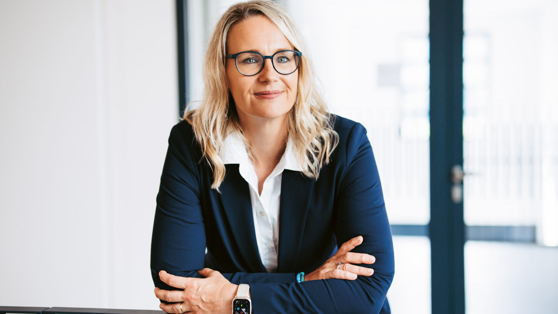 Eine Frau mit blondem Haar und Brille, die einen marineblauen Blazer trägt, steht mit verschränkten Armen in einem Haus und lächelt selbstbewusst.