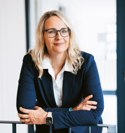 Eine Frau mit blondem Haar und Brille, die einen marineblauen Blazer trägt, steht mit verschränkten Armen in einem Haus und lächelt selbstbewusst.