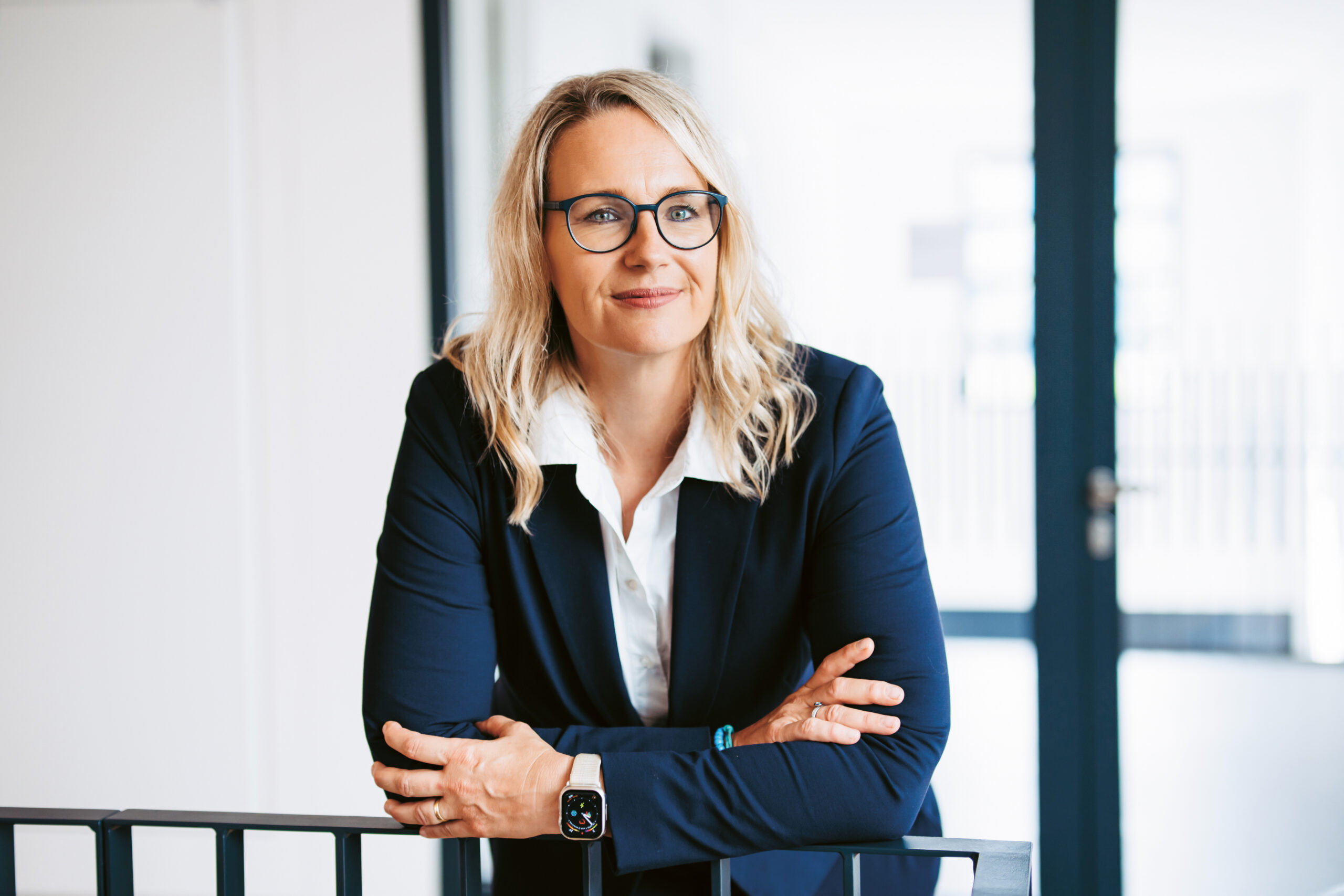 Eine Frau mit blondem Haar und Brille, die einen marineblauen Blazer trägt, steht mit verschränkten Armen in einem Haus und lächelt selbstbewusst.