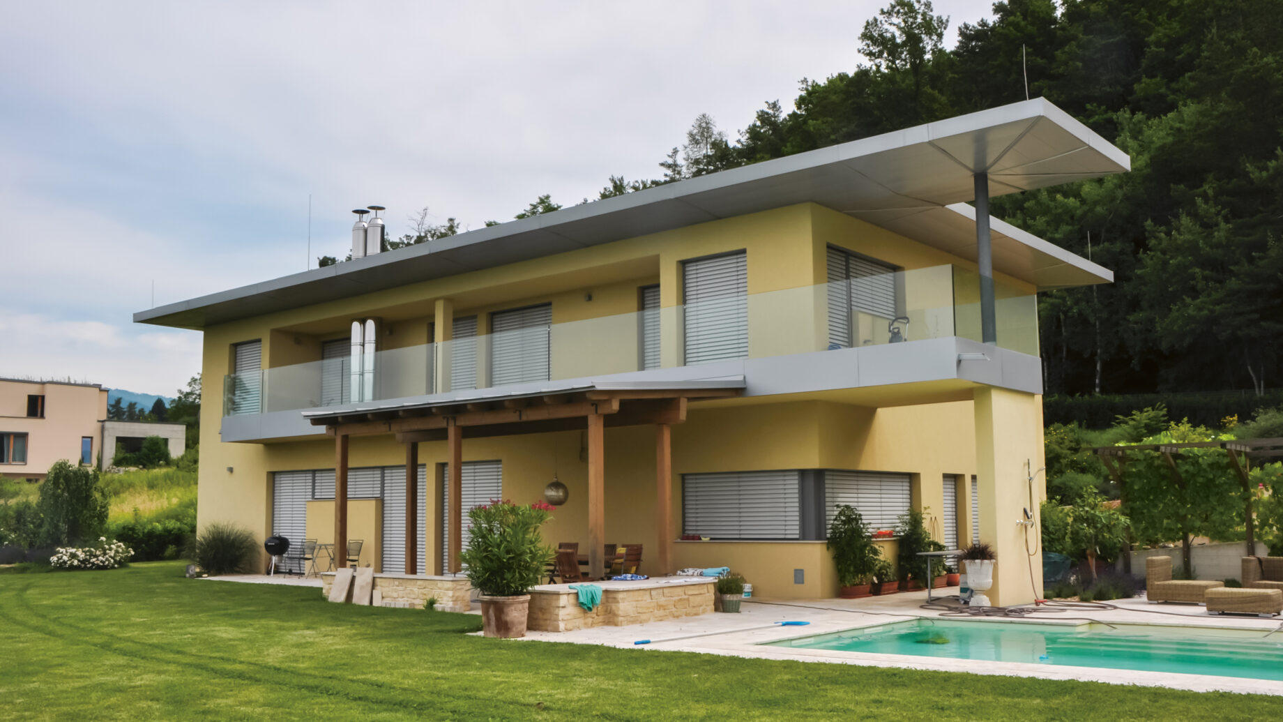 Modernes zweistöckiges gelbes Haus mit großem Balkon, Glasgeländer und einem Pool im Garten, umgeben von grünem Rasen und Bäumen.