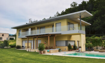 Modernes zweistöckiges gelbes Haus mit großem Balkon, Glasgeländer und einem Pool im Garten, umgeben von grünem Rasen und Bäumen.