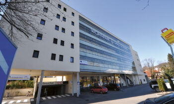 Ein modernes mehrstöckiges Gebäude mit der Aufschrift "Wohnpark Gaisberg", mit Geschäften im Erdgeschoss, parkenden Autos und einem strahlend blauen Himmel.