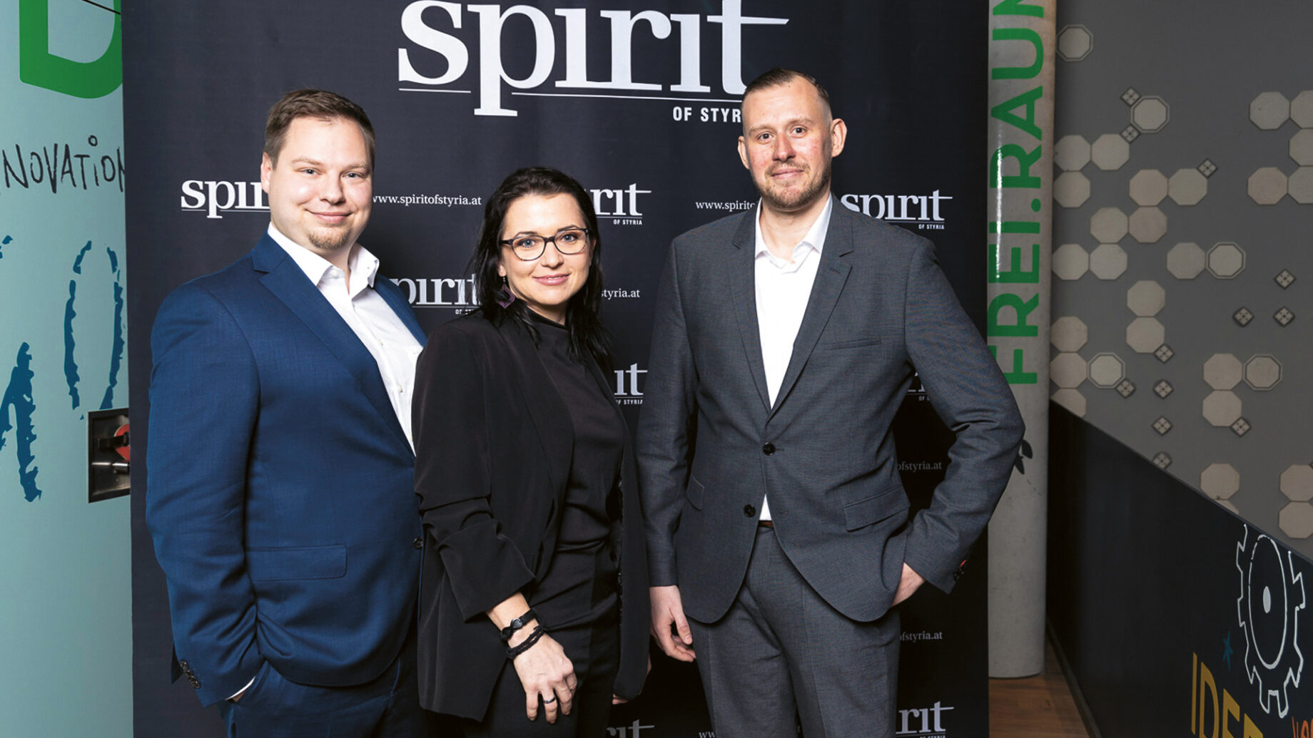Drei Personen in Business-Kleidung stehen lächelnd vor einem schwarzen Hintergrund mit dem "spirit of styria"-Logo bei einer professionellen Veranstaltung.