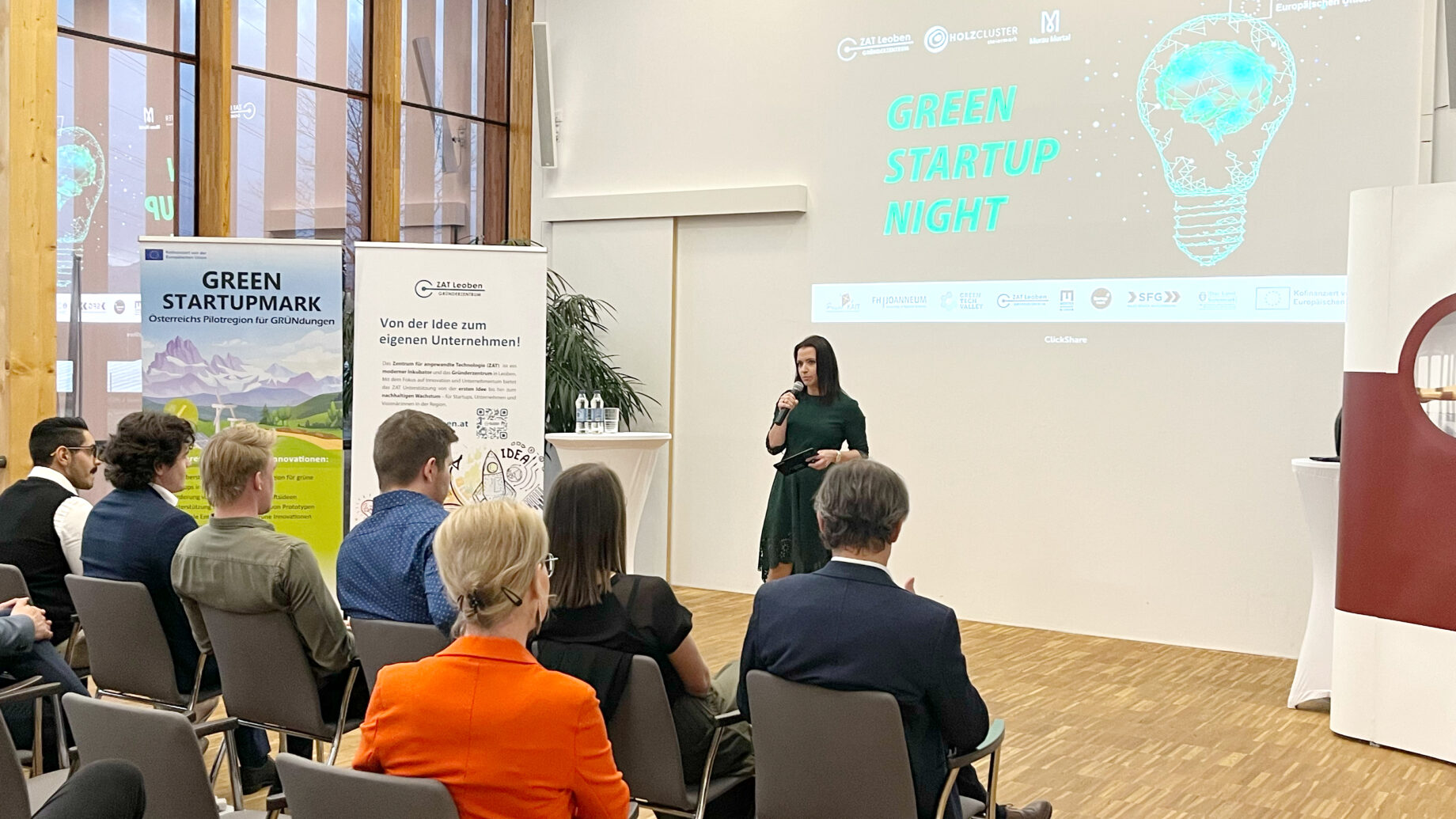 Eine Frau spricht auf einer Indoor-Veranstaltung namens "Green Startup Night" zu den Zuhörern, während im Hintergrund Banner und eine Präsentation zu sehen sind.