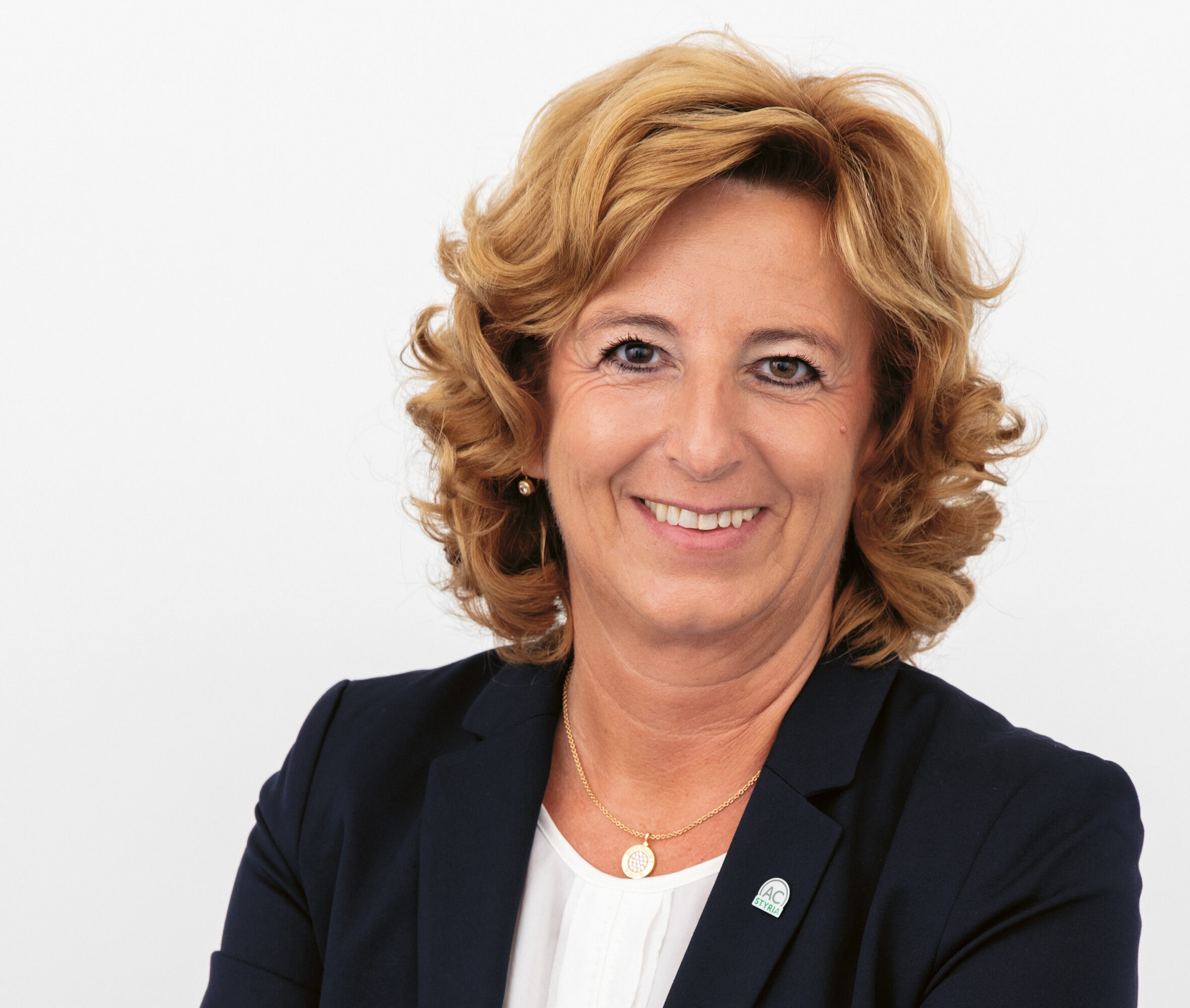 Lächelnde Frau mit lockigem blondem Haar, die einen marineblauen Blazer und eine weiße Bluse trägt, steht mit verschränkten Armen vor einem schlichten weißen Hintergrund.