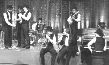 Eine Jazzband in Westen und Bowlerhüten spielt auf der Bühne mit verschiedenen Instrumenten, darunter Klavier, Saxophon, Posaune und Schlagzeug.